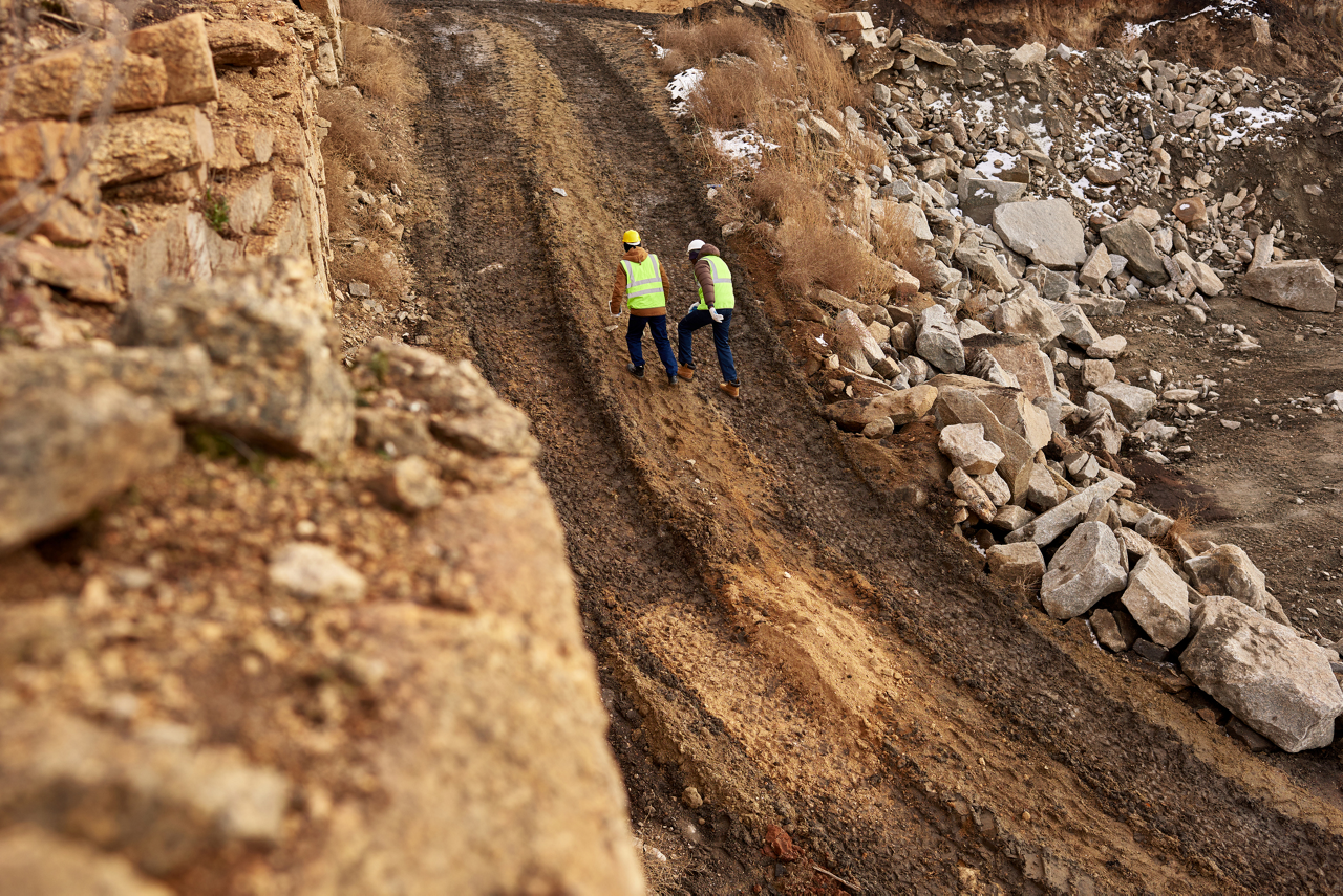 Two construction works follow a trail of large tire tracks.