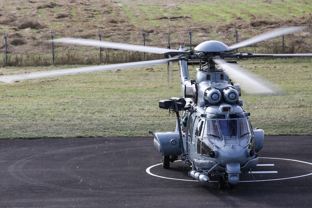 An Airbus H225M military helicopter is shown on a helipad with its main and tail rotor systems turning. 