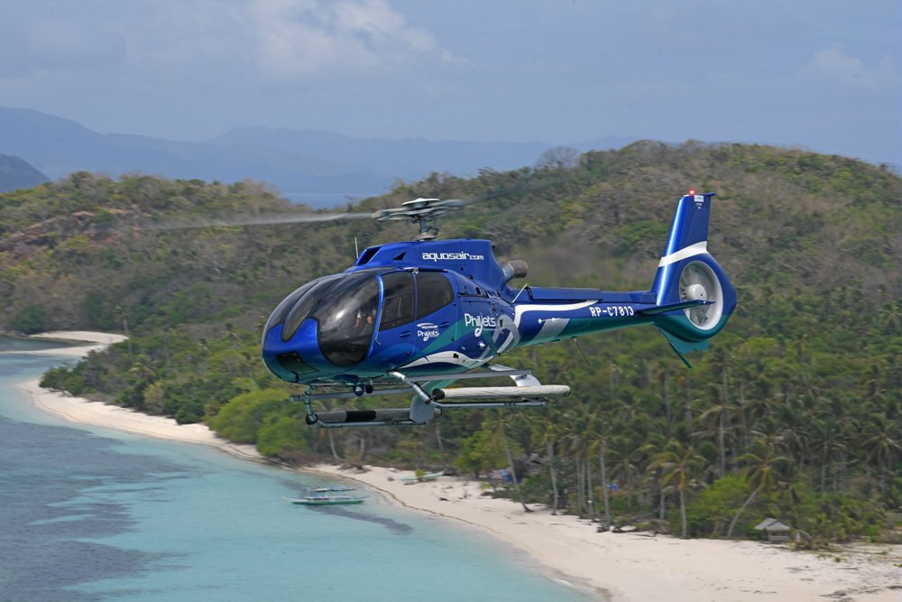 A Philjets-operated H130 helicopter flies over a picturesque beach setting 