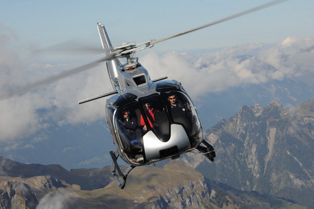 Head-on view of an in-flight Airbus H130 helicopter operating above mountainous terrain. 