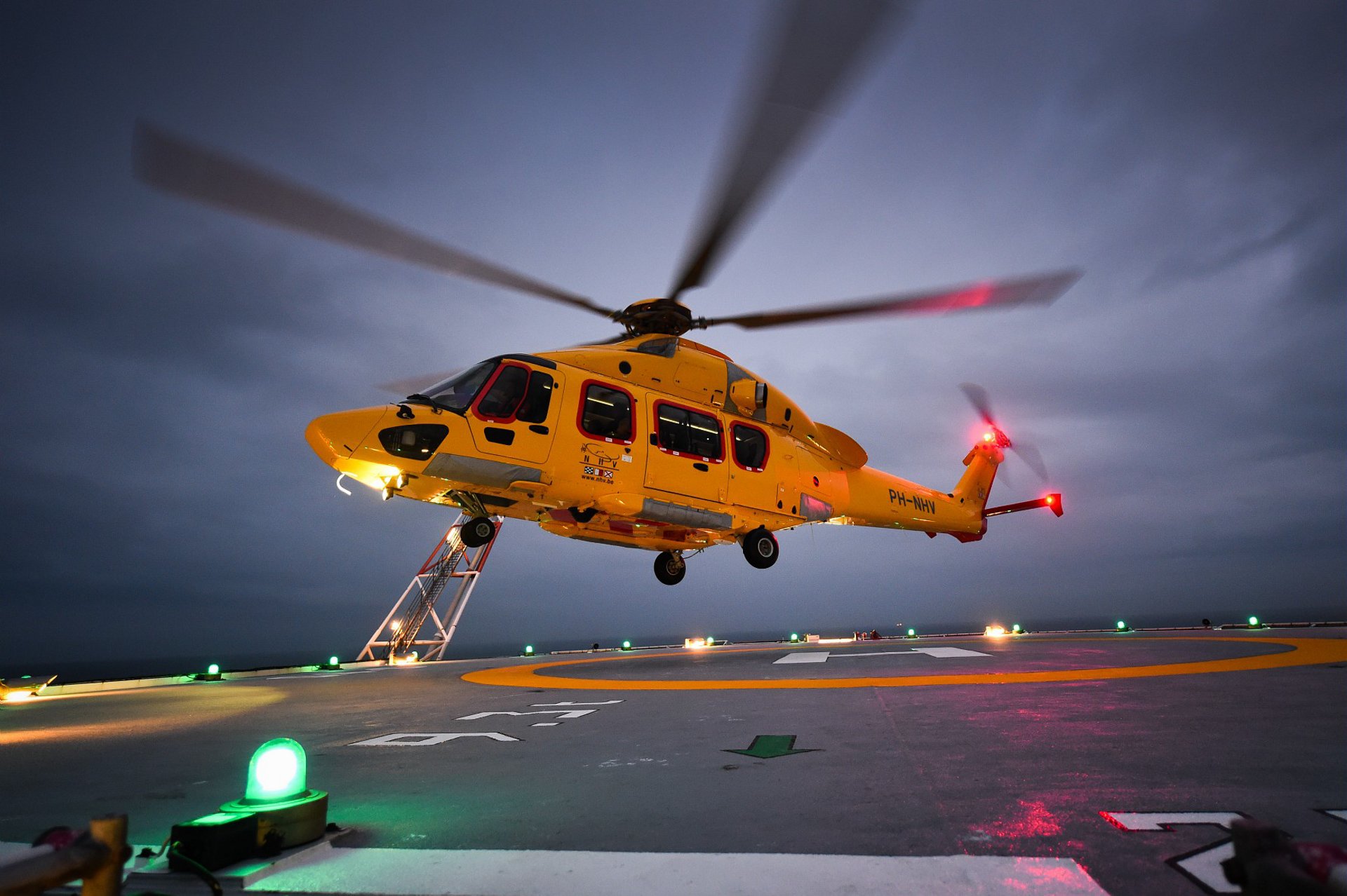 An Airbus H175 helicopter hovers over a helipad with dark skies above.
