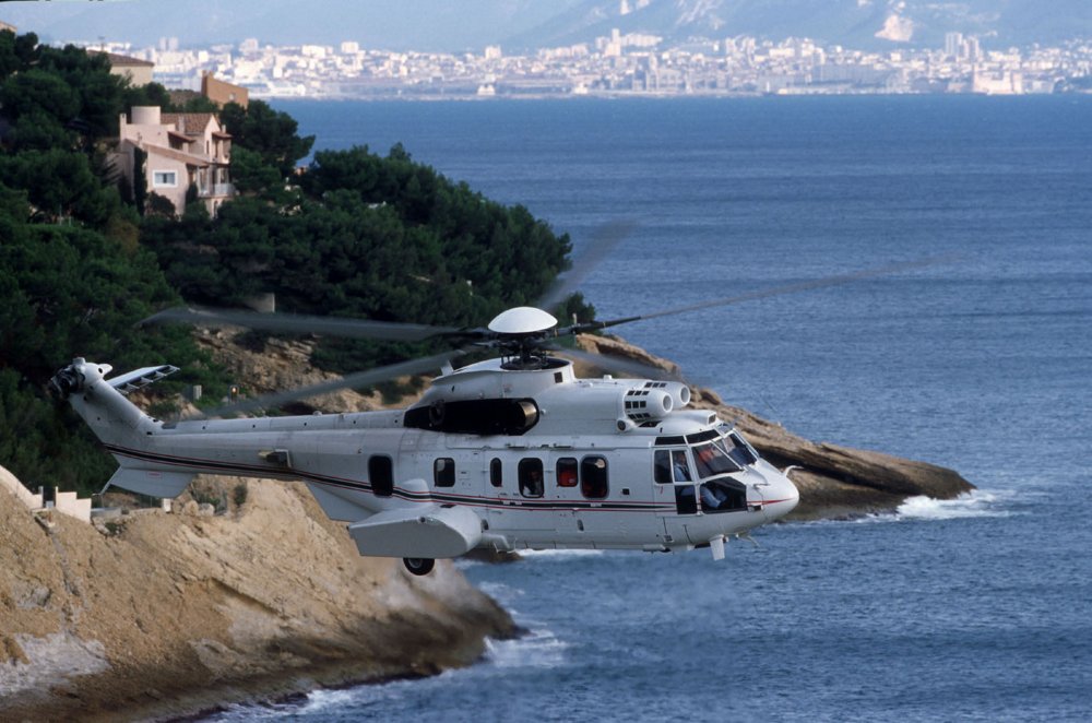 An Airbus H225 helicopter configured for VIP transport flies over a coastal area. 
