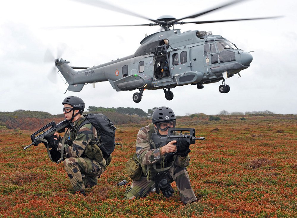 An Airbus H225M military helicopter takes off with two crouching soldiers shown in the foreground.  