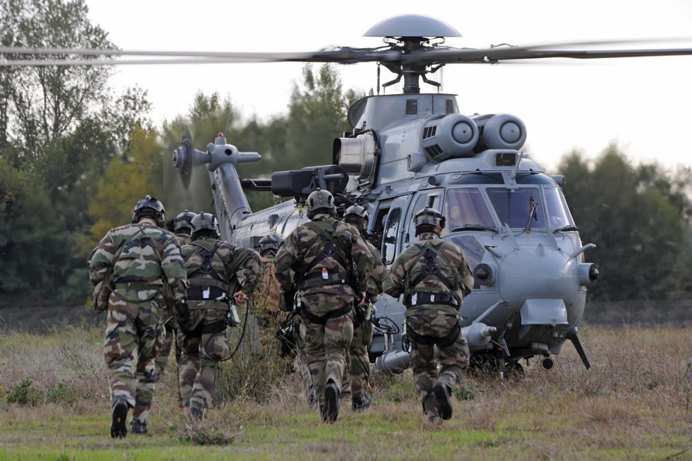 A group of military personnel advance toward an on-ground Airbus H225 military helicopter. 