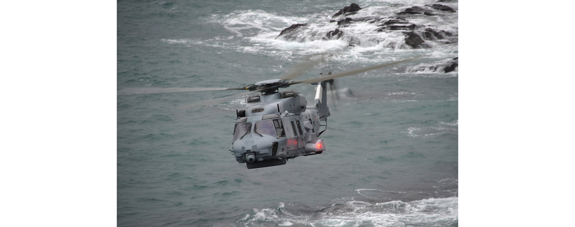 Head-on view of an NH90 NFH military helicopter flying over a coastal area. 