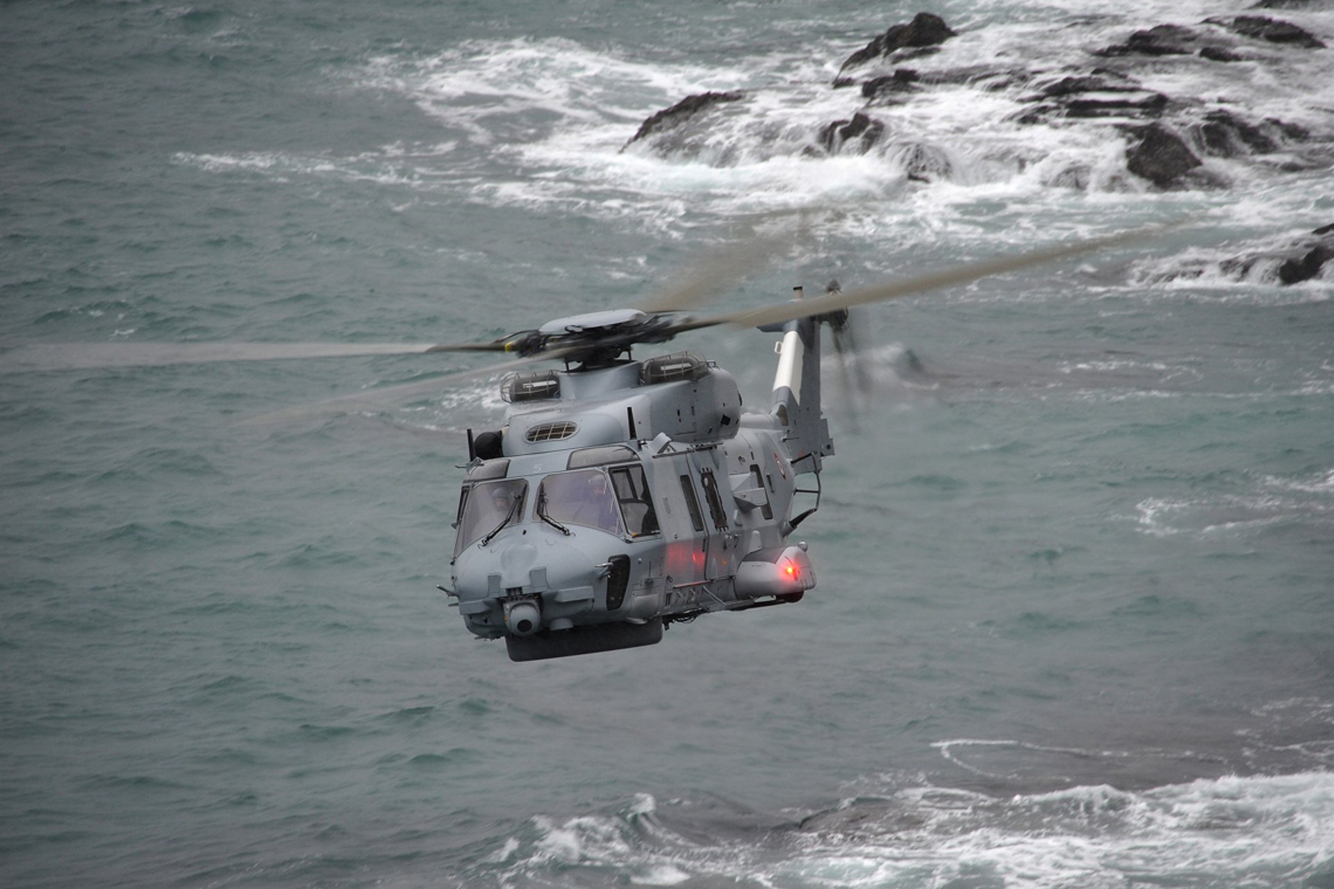 Head-on view of an NH90 NFH military helicopter flying over a coastal area.