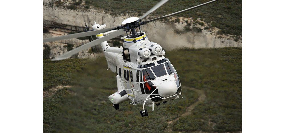 An Airbus H225 helicopter flies over varied terrain. 