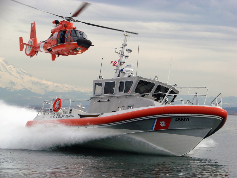 An in-flight Airbus AS565 MBe helicopter delivered to the U.S. Coast Guard trails a speeding Coast Guard vessel. 