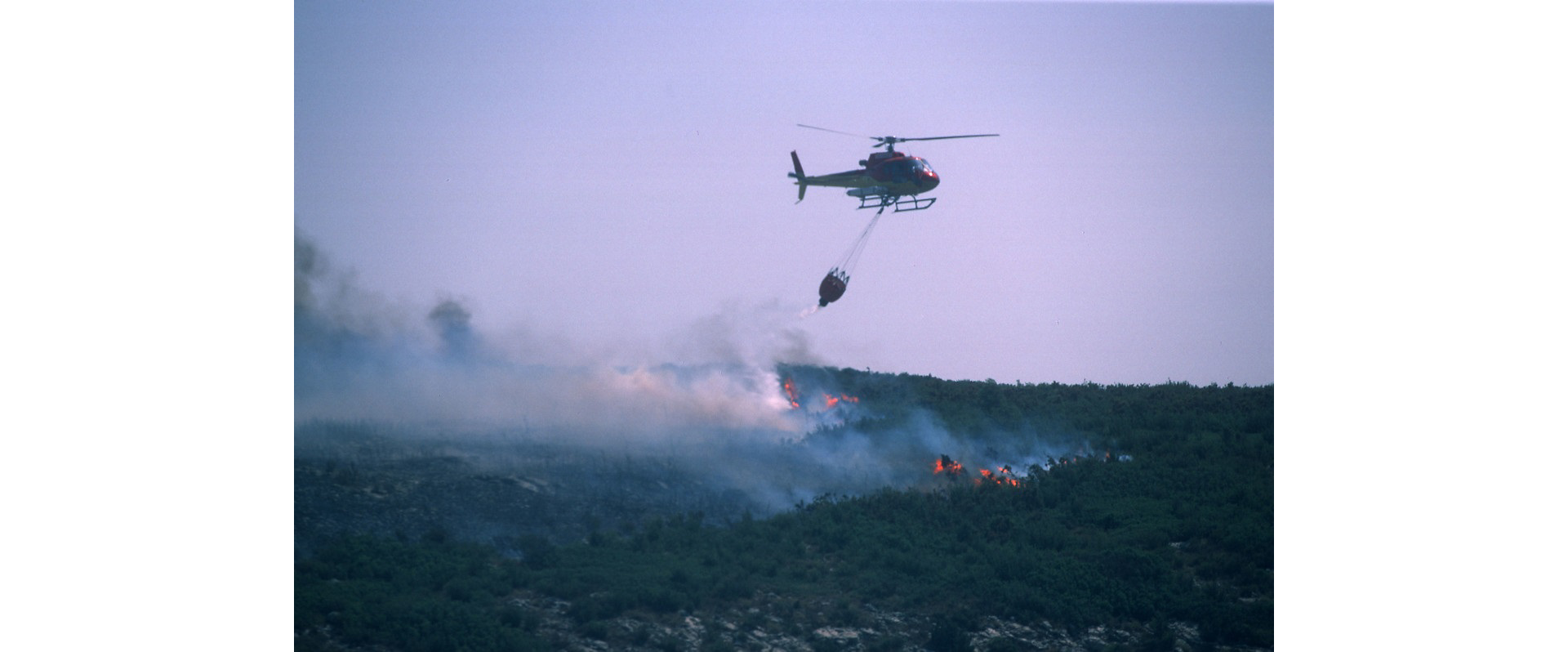 A helicopter fights a wildfire with deployment of a bucket-type water dispersal system.