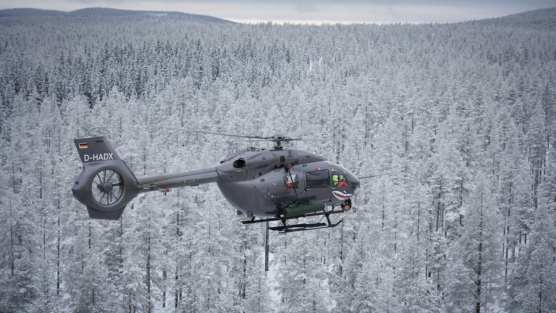 An Airbus H145M military helicopter flies over a wooded area covered with snow. 