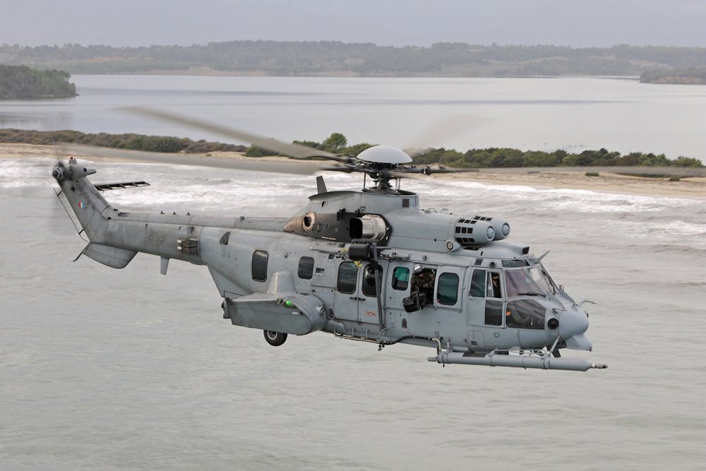 An H225M military helicopter flies over a coastal setting.  