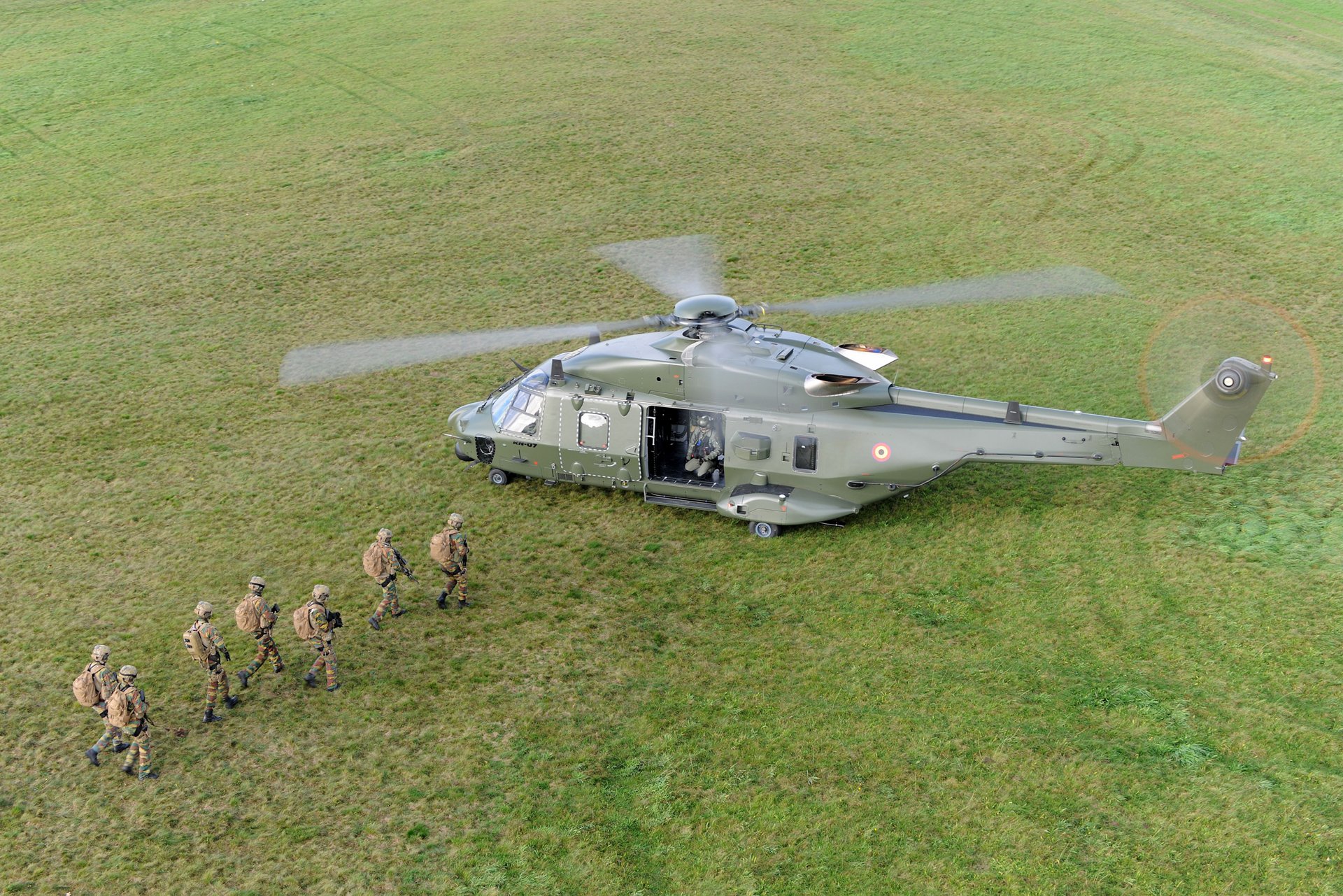 A group of seven military troops advance toward an on-ground NH90 helicopter. 