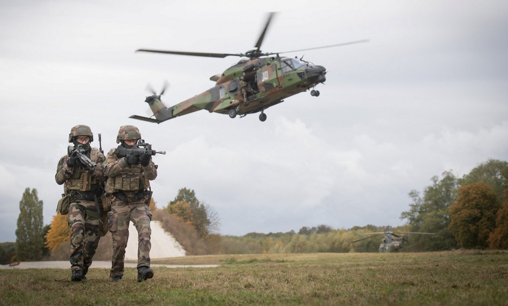 两名作战部队前进，一架NH90直升机在背景中起飞。